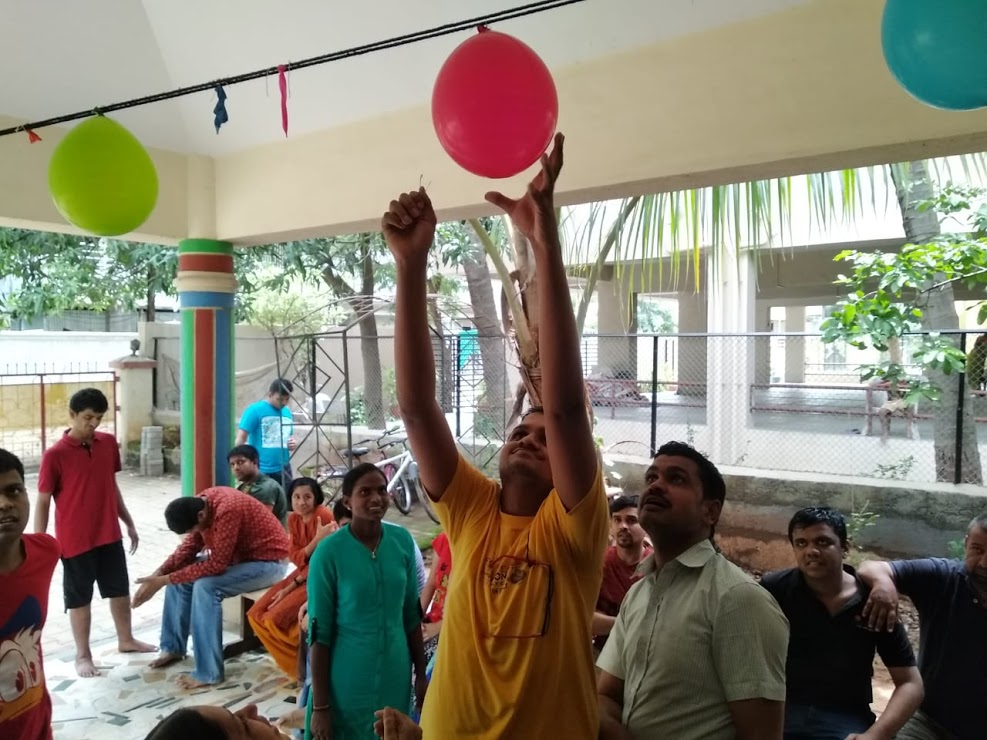 A participant in a yellow t-shirt reaches up joyfully to tap a balloon during a group celebration activity
